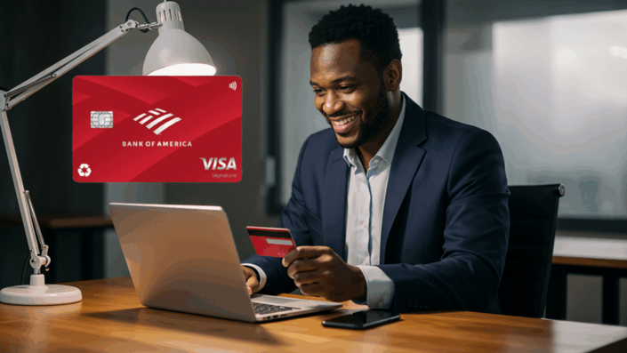 Hombre feliz usando una tarjeta de crédito Bank of America Customized Cash Rewards mientras trabaja en una oficina moderna con luz artificial y una computadora portátil.