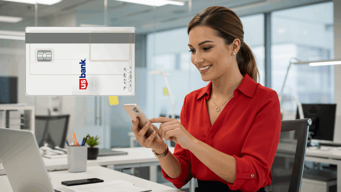 Mujer feliz trabajando en una oficina moderna, usando un teléfono móvil con una tarjeta de crédito US Bank Shield Visa visible en la parte superior izquierda.