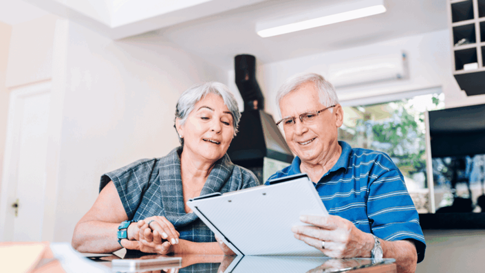 Pareja de adultos mayores sonriendo mientras revisan documentos en una tableta, planificando su Ahorro para la jubilación en la mesa de su casa.