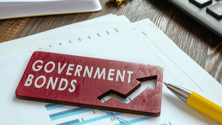 Placa de madera roja con las palabras "GOVERNMENT BONDS" (Bonos Gubernamentales) y una flecha ascendente recortada, sobre papeles con gráficos financieros y un bolígrafo amarillo, simbolizando la inversión en bonos del gobierno.