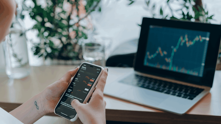 Persona revisando gráficos de acciones en un smartphone, con una laptop mostrando datos similares al fondo, representando la decisión de ahorro o inversión en el mercado financiero.