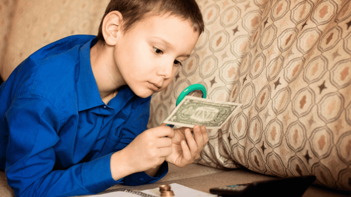 Niño en camisa azul examinando un billete de un dólar con una lupa, un ejemplo de Educación financiera para niños y el interés por el valor del dinero.