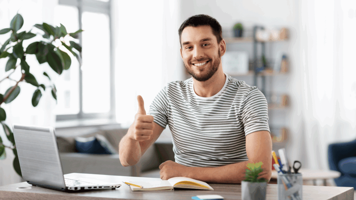Hombre freelancer sonriente sentado en su escritorio con una laptop y un cuaderno, haciendo un gesto de pulgar hacia arriba, simbolizando el éxito en la gestión de finanzas para freelancer.