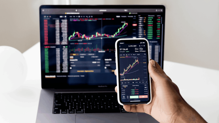 Persona sosteniendo un teléfono inteligente que muestra un gráfico financiero, frente a una laptop con una pantalla de gráficos y datos bursátiles, representando el seguimiento e inversión en fondos indexados en EE. UU.