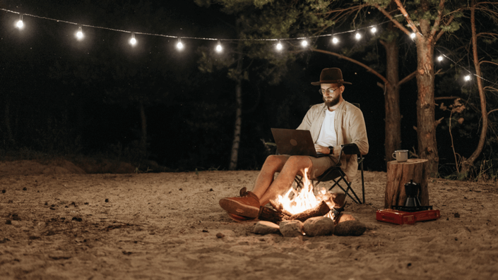 Hombre freelancer con sombrero y gafas trabajando en su laptop junto a una fogata por la noche, bajo una guirnalda de luces, representando el estilo de vida flexible del freelancer.