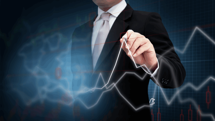 A businessman in a black suit and light pink tie is drawing a white line on a graph, analyzing trends related to Stock market volatility. The graph has a blue background with a grid pattern and shows a stock market chart with red and green candlesticks.
