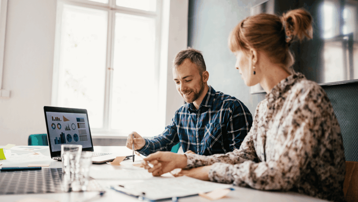 Dos personas trabajando juntas frente a una computadora con gráficos financieros, discutiendo estrategias de inversiones para alcanzar objetivos económicos.