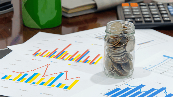 Frasco lleno de monedas sobre papeles con gráficos de barras y líneas, simbolizando el análisis y seguimiento de inversiones para maximizar rendimientos.