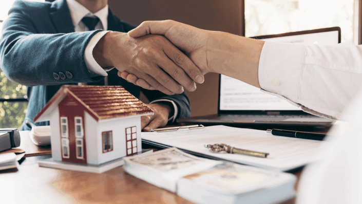 Apretón de manos entre dos personas en oficina con maqueta de casa y documentos, representando acuerdos y negociaciones en inversiones inmobiliarias