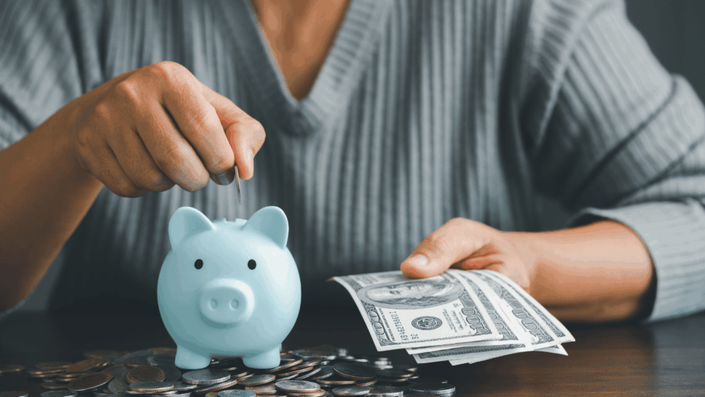Persona colocando una moneda en una alcancía azul claro, rodeada de monedas y sosteniendo billetes de dólar, representando el concepto de inversiones seguras.