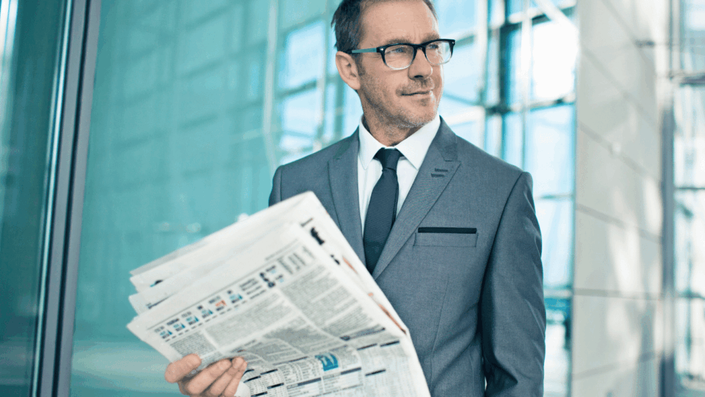 A man in a grey suit and glasses is holding a stack of newspapers, staying informed about factors affecting Stock market volatility, in front of a modern building with glass windows.