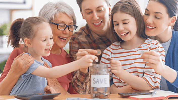 Familia de varias generaciones sonriendo mientras enseñan a los niños a ahorrar dinero metiendo monedas en un frasco etiquetado como "SAVE", destacando la importancia de la priorización del ahorro en el presupuesto familiar.