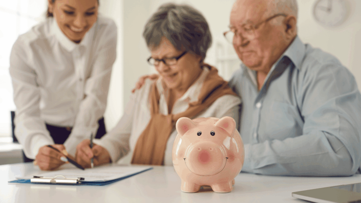 Pareja de ancianos y una agente financiera firmando documentos mientras un cochinito de ahorro destaca, reflejando cómo un seguro de vida puede ser parte esencial de la planificación financiera en la tercera edad.