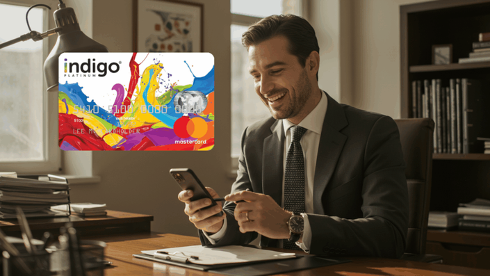 Businessman in a suit smiling while using his smartphone in a professional office, with an enlarged image of the Indigo Platinum Mastercard displayed beside him.