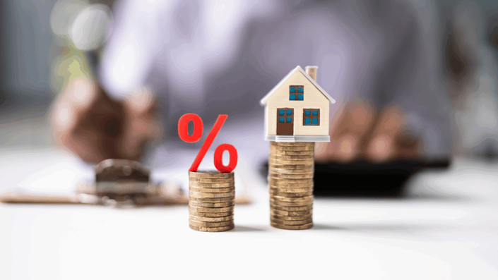 Símbolo de porcentaje rojo sobre una pila de monedas junto a una casa miniatura sobre otra pila de monedas, ilustrando el concepto de la tasa de interés en el sector inmobiliario o inversiones. Una persona calcula en el fondo.