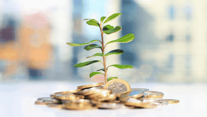 Pequeña planta verde brotando de un montón de monedas de euro, con un fondo urbano desenfocado, representando el crecimiento y las nuevas Tendencias de inversión.