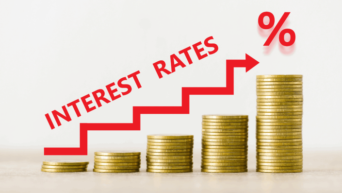 Gráfico de barras con pilas de monedas doradas de alturas crecientes de izquierda a derecha, con una flecha roja ascendente y el texto 'INTEREST RATES' y un símbolo '%' sobre la pila más alta, representando el aumento de la tasa de interés.