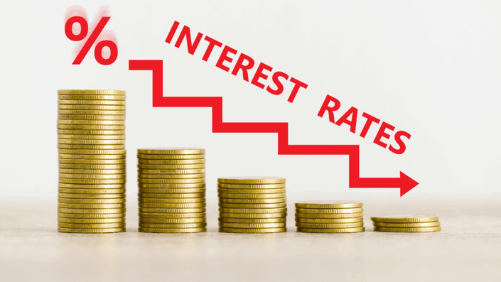 Gráfico de barras con pilas de monedas doradas de alturas decrecientes de izquierda a derecha, con una flecha roja descendente y el texto 'INTEREST RATES' y un símbolo '%' sobre la pila más alta, representando la caída de la tasa de interés.